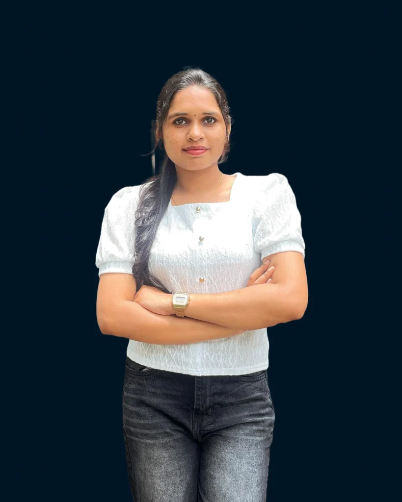 Ajuna, a freelance digital marketing analyst in Kozhikode with medium dark skin and long dark brown hair in a side ponytail stands facing the camera with her arms crossed. She wears a white short sleeved textured top with a square neckline and three gold buttons, dark black jeans, a gold digital watch on her left wrist, small gold earrings, a thin gold necklace, and a bindi on her forehead. She has a slight smile. The background is solid dark navy blue.​​​​​​​​​​​​​​​​​​​​​​​​​​​​​​​​​​​​​​​​​​​​​​​​​​